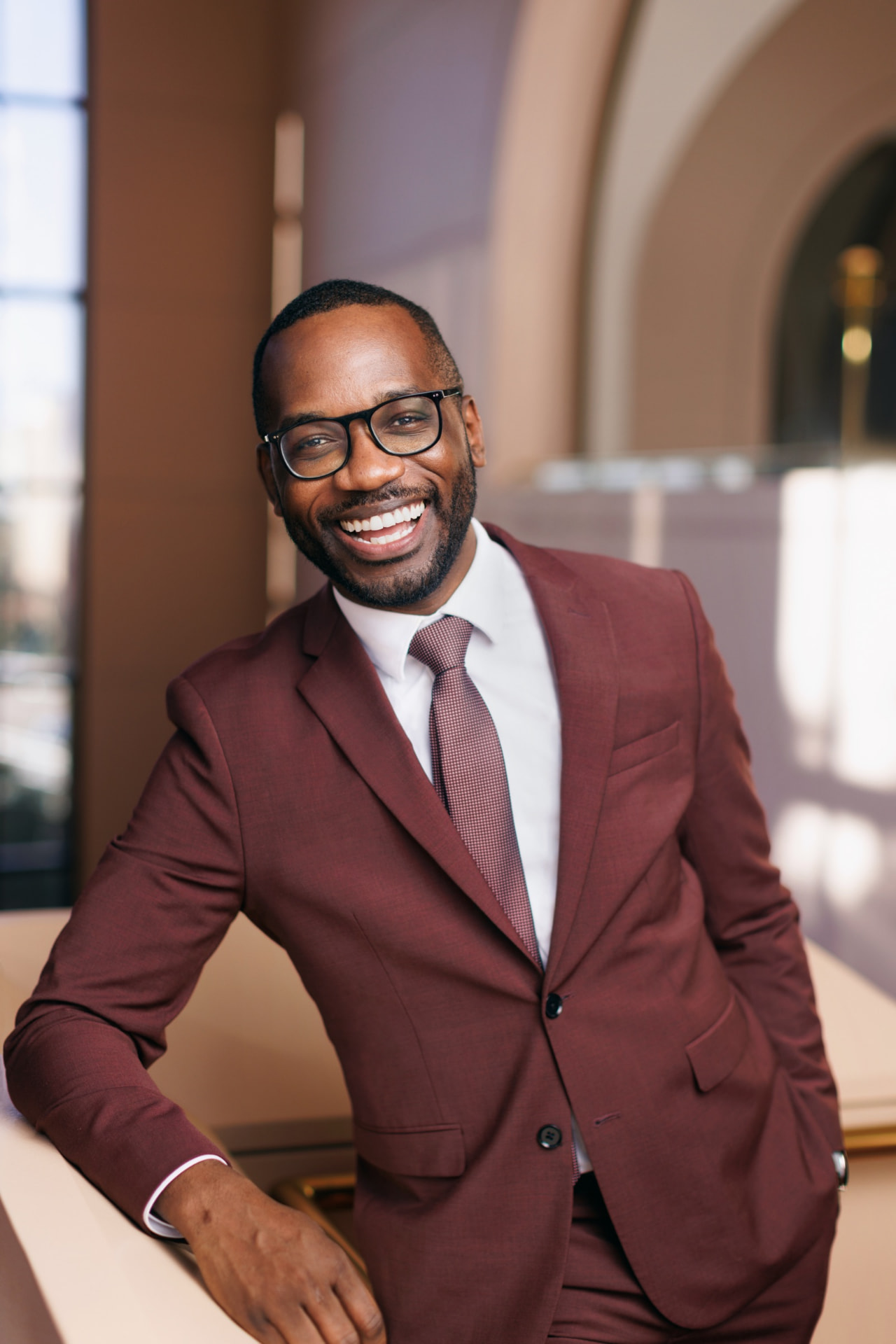 Joel Thompson in a maroon suit, smiling warmly and leaning on a railing in a sunlit stone interior.