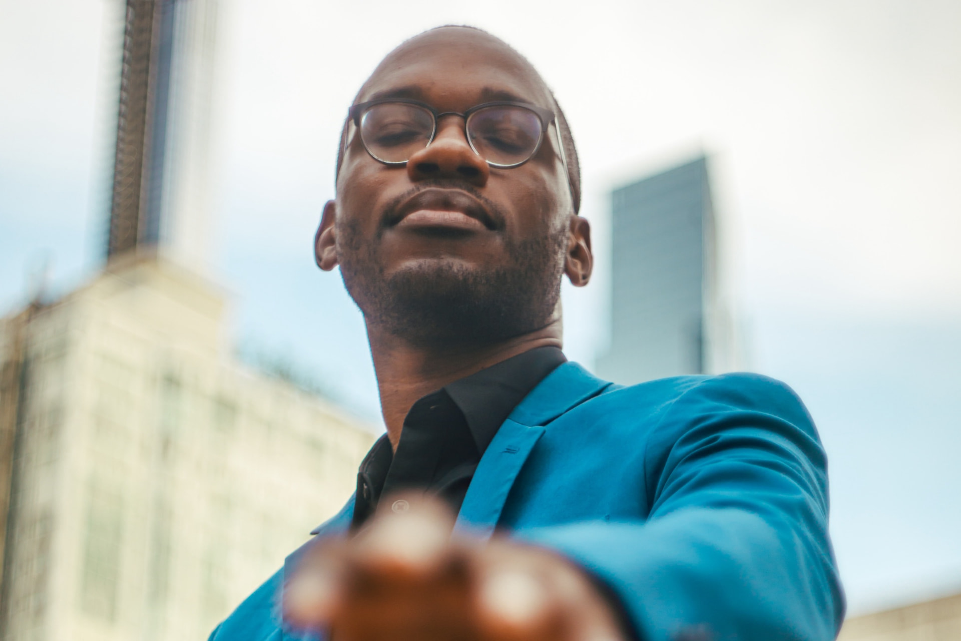 Joel Thompson in a teal-blue suit, photographed from a low angle, one hand reaching toward the camera, city towers behind him.