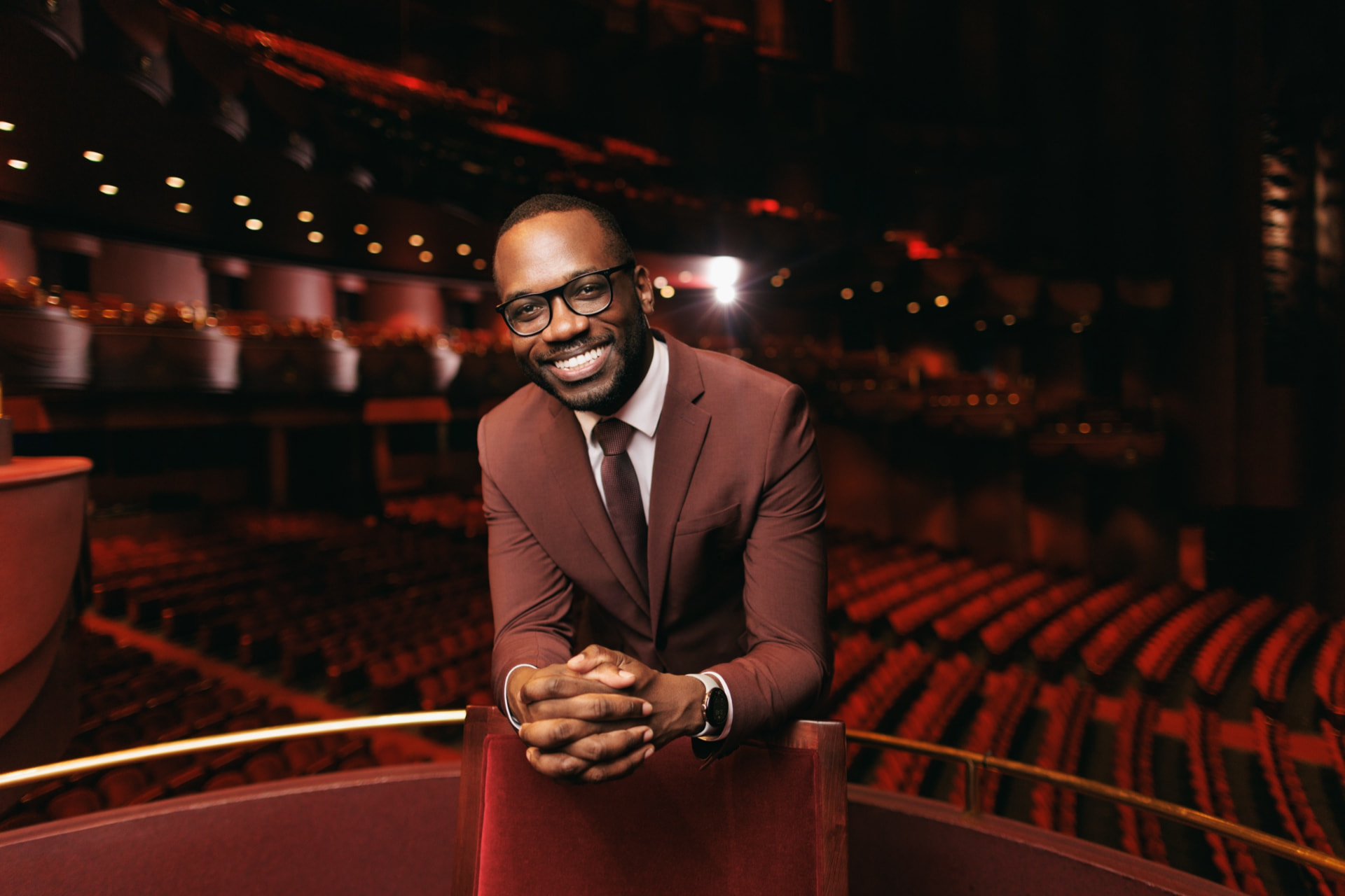 Composer Joel Thompson in a maroon suit, hands clasped on a red velvet seat inside a softly lit, empty opera house.