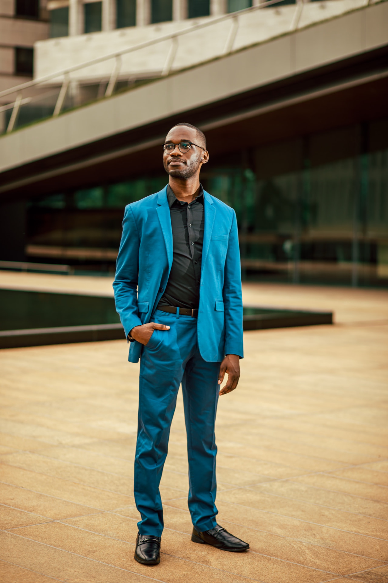 Joel Thompson standing full-length in a blue suit, one hand in his pocket, on a sunlit plaza of stone and glass.