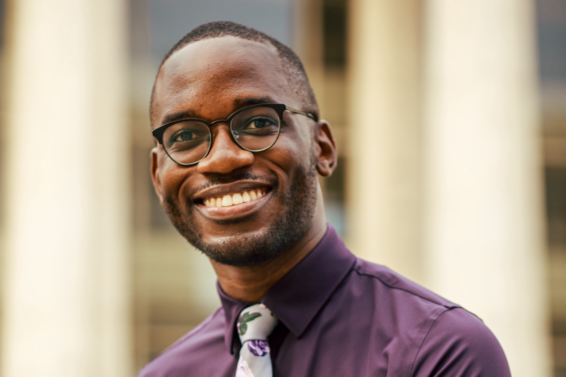 Joel Thompson in an aubergine shirt and floral tie, smiling warmly before a blurred colonnade.